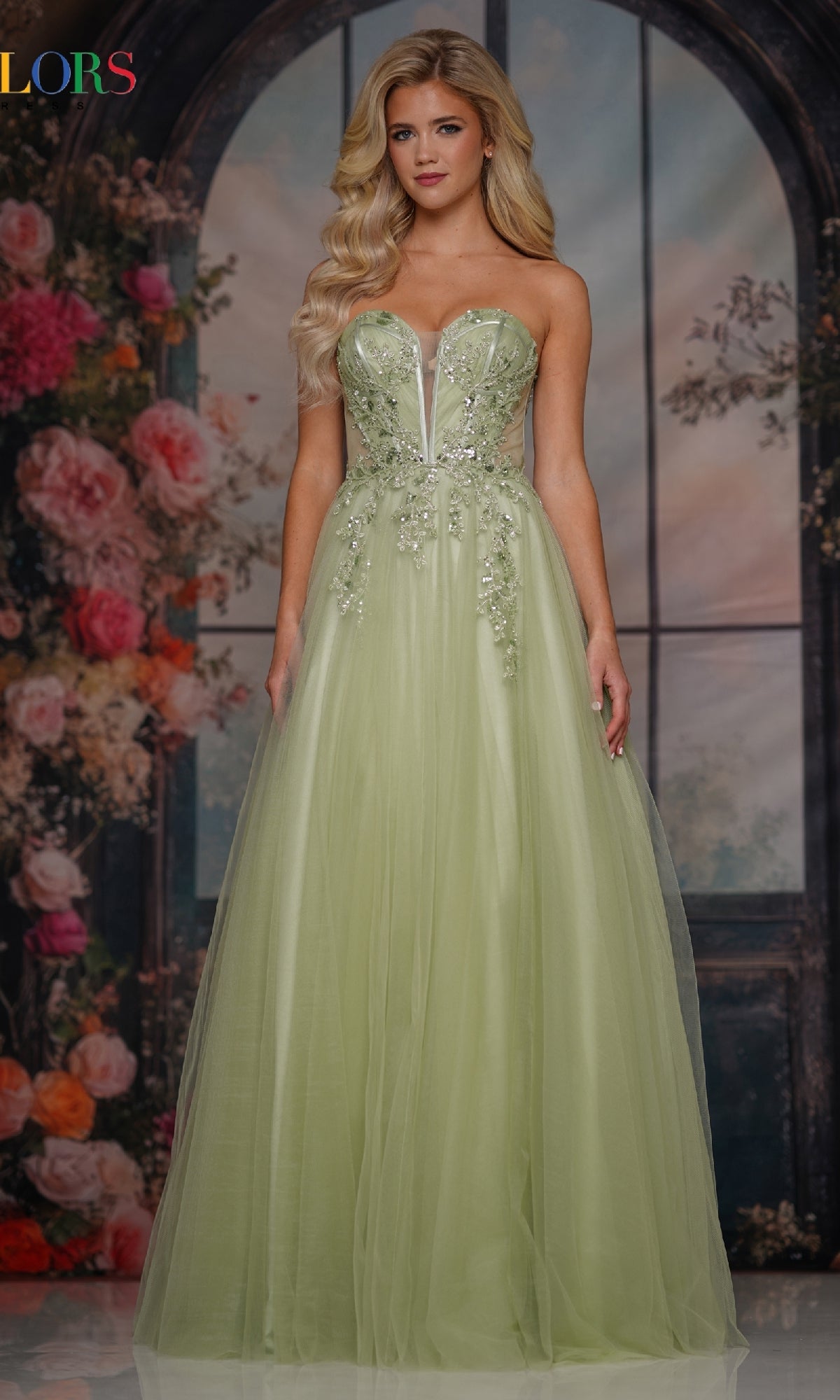 A woman wears the Strapless Long Corset Prom Dress: Colors Dress 3923 in light green, featuring a floral-embellished bodice, standing before a floral and arched window backdrop.